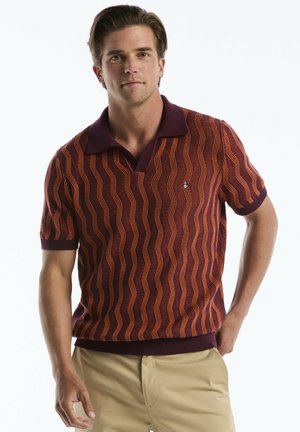 Mann trägt einen kurzärmeligen Pullover mit weinrotem und orangefarbenem Muster und beigen Hosen, steht mit einer Hand in der Tasche.