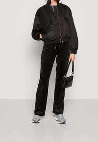 Blouson aviateur en satin noir avec fermeture éclair, poches latérales et poignets côtelés, associé à un pantalon en velours noir et des baskets grises.