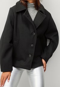 Veste en laine noire mélangée avec un col, quatre boutons et des poches latérales. Les manches sont légèrement oversize, associées à un pantalon argenté.