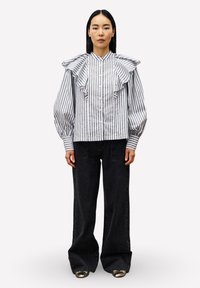 Vrouw in een wit met zwart verticaal gestreepte blouse met rimpel details op de schouders en zwarte wijde broek met metallic schoenen.
