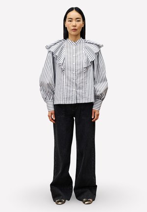 Vrouw in een wit met zwart verticaal gestreepte blouse met rimpel details op de schouders en zwarte wijde broek met metallic schoenen.
