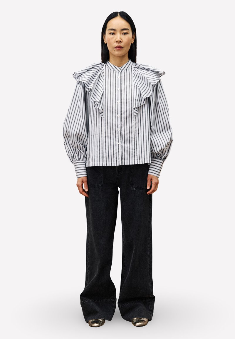Vrouw in een wit met zwart verticaal gestreepte blouse met rimpel details op de schouders en zwarte wijde broek met metallic schoenen.