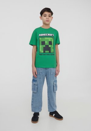 T-shirt verde in cotone con grafica di un Creeper di Minecraft, abbinato a pantaloni cargo blu chiaro e sneakers nere con accenti bianchi.