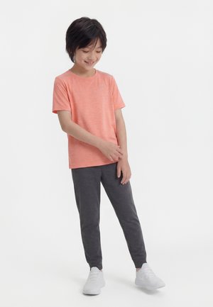 Niño con una camiseta coral de manga corta, pantalones joggers gris oscuro y zapatillas blancas, de pie y mirando hacia abajo con una sonrisa.
