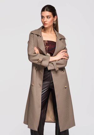 Mujer con el cabello recogido, que lleva pendientes grandes, un abrigo tipo gabardina con estampado, una blusa estampada y pantalones de cuero negro, de pie con los brazos cruzados.