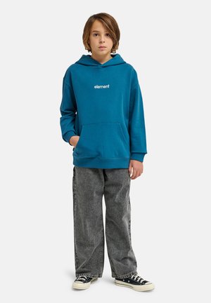 Kind mit mittellangen Haaren, das einen blauen "Element" Hoodie, graue lockere Jeans und schwarze flache Sneakers trägt und mit einer Hand in der Tasche steht.