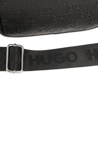 Sac à épaule noir texturé avec logo "HUGO" en relief, doté d'une sangle plate avec une boucle en métal argenté et des détails de couture.