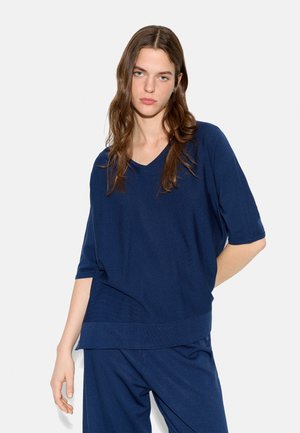 Młoda kobieta z długimi brązowymi włosami, ubrana w luźny granatowy top z dekoltem w serek oraz dopasowane spodnie na jednolitym tle.