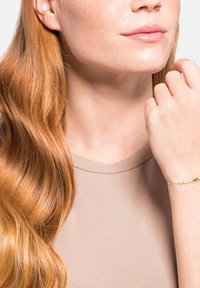 Pulsera de oro con piedras preciosas de colores, incluyendo púrpura y verde. Llevada en la muñeca de una mujer, con una camisa neutra y cabello largo, ondulado y castaño.