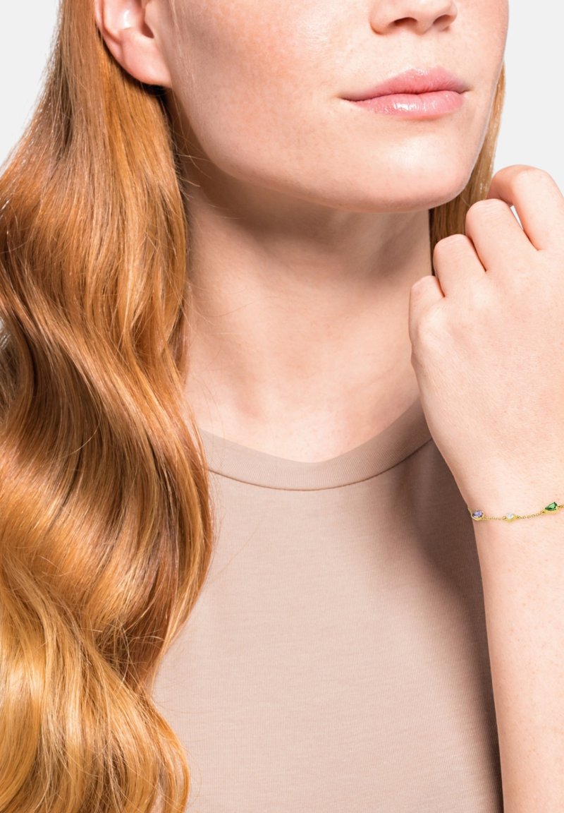 Pulsera de oro con piedras preciosas de colores, incluyendo púrpura y verde. Llevada en la muñeca de una mujer, con una camisa neutra y cabello largo, ondulado y castaño.