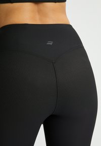 Las mallas negras de canalé presentan una cintura alta y un logo reflectante en la parte posterior, exhibiendo un acabado suave y texturizado.