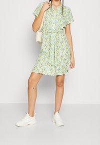 Abito senza maniche con motivo floreale verde e blu, cincherato in vita da una cintura. Abbinato a sneakers bianche e una borsa color crema.