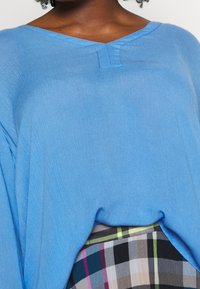 Blauw V-hals top van een lichte, gestructureerde stof. Heeft een losse pasvorm en zichtbare naden. Gekleed met een meerkleurige geruite rok.