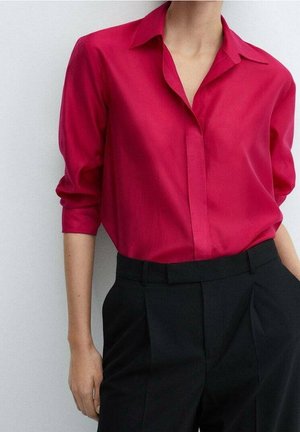 Personne portant une chemise boutonnée à manches longues rouge vif, rentrée dans un pantalon noir taille haute, sur un fond uni.