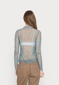 Blusa transparente de mangas longas com padrão verde em ondas, gola alta e design ajustado. Complementada por uma camada inferior azul clara. Calças castanhas.