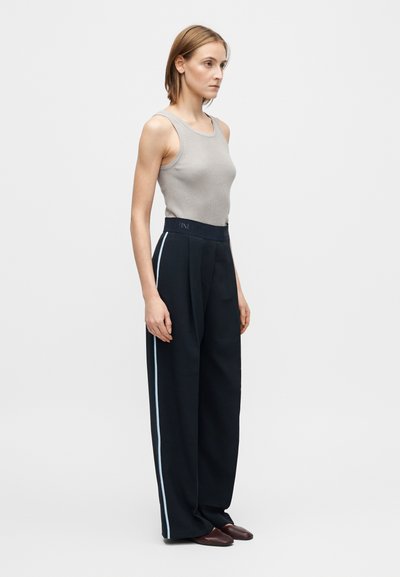 Grijze geribbelde tanktop gecombineerd met zwarte broek met wijde pijpen en een lichtblauwe zijstreep. Het model staat in profiel en draagt donkere schoenen.