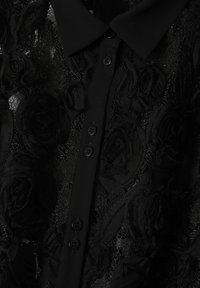 Blusa nera con bottoni, realizzata in un tessuto trasparente strutturato con motivi floreali neri in rilievo. Design con colletto e bottoni neri.