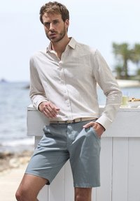Hellbeige gestreifte Langarmbluse kombiniert mit blauen Shorts, versehen mit einem geflochtenen Gürtel, vor einem Strandhintergrund.