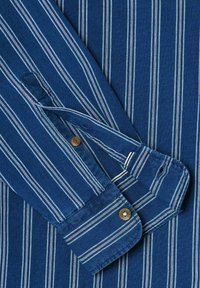 Stripet blå skjorte med vertikale hvide og røde striber, knapdetalje ved manchet og en blød tekstur.