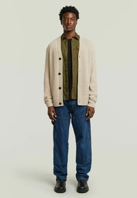 Homme debout portant un cardigan beige à boutons, une chemise vert olive par-dessus un top noir, un jean bleu et des chaussures noires, sur fond clair.