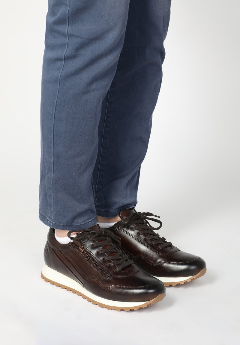 Braune Leder-Sneaker mit strukturiertem Finish, ausgestattet mit einer weißen Zwischensohle, brauner Außensohle und schwarzen Schnürsenkeln. Kombiniert mit dunkelbblauen Hosen.