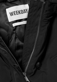 Jachetă neagră cu exterior neted, având un fermoar argintiu, etichetă cu inscripția "WEEKDAY" și un detaliu cu buton pe lateral.