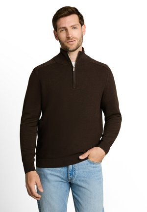 Man met een donkerbruine vest met rits en lichtblauwe spijkerbroek, één hand in de zak, staand tegen een effen witte achtergrond.
