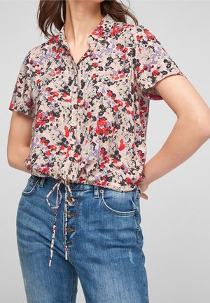 Femme portant un chemisier à manches courtes à fleurs violettes, rouges et noires, assorti à un jean bleu taille haute.