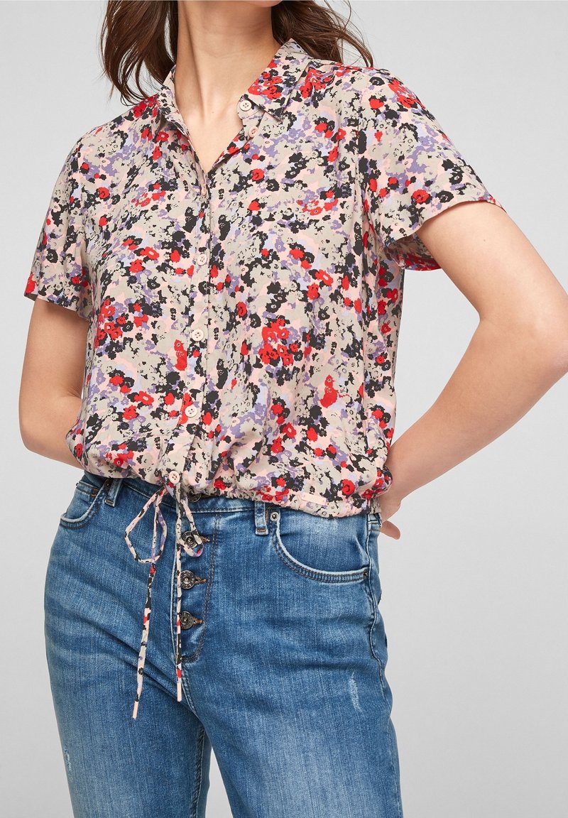 Femme portant un chemisier à manches courtes à fleurs violettes, rouges et noires, assorti à un jean bleu taille haute.