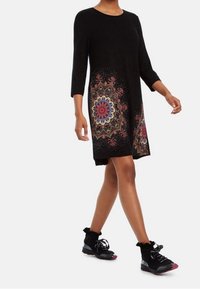 Svart klänning med trekvarts ärmar som har färgglada mandala-mönster längs fållen, parat med svarta träningssko.