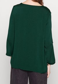 Kvinna i en lös passform, mörkgrön långärmad blus och mörkgrå byxor, sedd bakifrån framför en enkel bakgrund.