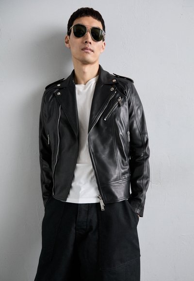 Giubbotto da biker in pelle nera con tasche con zip, spalline, e un design aderente, abbinato a una t-shirt bianca e pantaloni scuri.