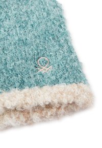 Tyrkysová chlupatá čepice s krémovým lemem, zdobená vyšívaným symboliem. Textura je měkká a chlupatá, s hrubým pleteným designem.