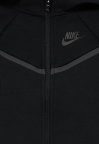 Felpa nera Nike con zip realizzata in tessuto morbido. Presenta una sottile striscia grigia e il logo sul petto, con un colletto alto e una cerniera liscia.