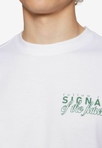 Witte katoenen t-shirt met een ronde nek, met groene tekst die zegt "volg het TEKEN van de toekomst" in verschillende letterstijl.