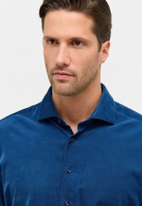 Camicia blu con bottoni, dallo stile classico, colletto a punta e sei bottoni scuri sul fronte. Dettagli o motivi limitati e poco visibili.