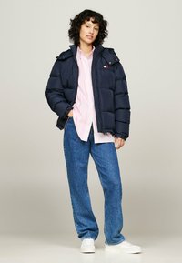Marine pufferjack met een capuchon, gestikte design, ritssluiting aan de voorkant en logoaccent. Gedragen over een roze gestreepte shirt en gecombineerd met een blauwe spijkerbroek.