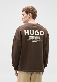 Maglietta marrone a maniche lunghe con la scritta "HUGO" e un logo vintage bianco sul retro, con una vestibilità comoda e una texture morbida.