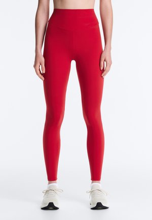 Femme portant des leggings rouges taille haute et des baskets blanches, debout avec les bras détendus le long du corps devant un fond blanc uni.