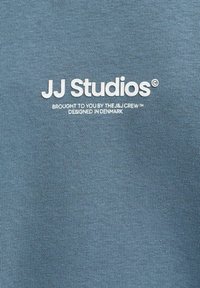 Μπλε ύφασμα με λευκό κείμενο που αναγράφει JJ Studios, προσφορά της ομάδας J&J, σχεδιασμένο στη Δανία.