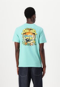 T-shirt de manga curta em turquesa com uma gráfica colorida nas costas, apresentando elementos de praia e florais, complementados por texto em destaque.