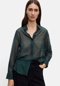 Zielona koszula z długim rękawem, półprzezroczysta, z zapięciem na guziki z przodu, kołnierzykiem i subtelną kropeczkowaną teksturą, w zestawieniu z ciemnymi spodniami.