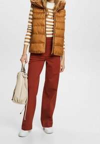Femme portant un gilet matelassé marron sur un haut rayé blanc et marron, un pantalon couleur rouille, des baskets blanches, tenant un sac à main beige.