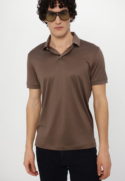 Brown polo shirt with short sleeves and a collar, featuring a subtle logo above the left side. Worn with dark pants and oversized sunglasses.