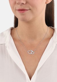Collar de plata con diseño de corazón y lazo, que presenta acentos brillantes. La cadena es delgada y delicada contra una camisa de color claro.