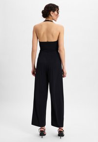 Schwarzer ärmelloser Jumpsuit mit weitem Bein und taillierter Oberkörper. Glattes Material mit Halterneck und einem kleinen Rückenausschnitt.