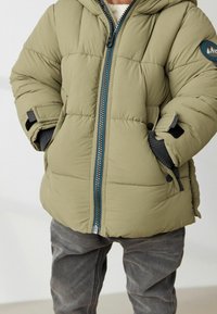 Chaqueta acolchada verde oliva con cierre de cremallera en la parte delantera, diseño acolchado, capucha y detalles en gris. Cuenta con un parche de logo redondo y puños ajustables.