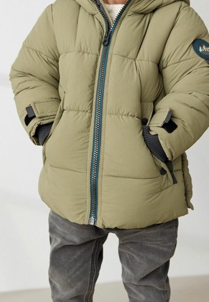 Giubbotto lungo verde oliva con zip frontale, design trapuntato, cappuccio e dettagli grigi. Presenta una toppa rotonda con logo e polsini regolabili.