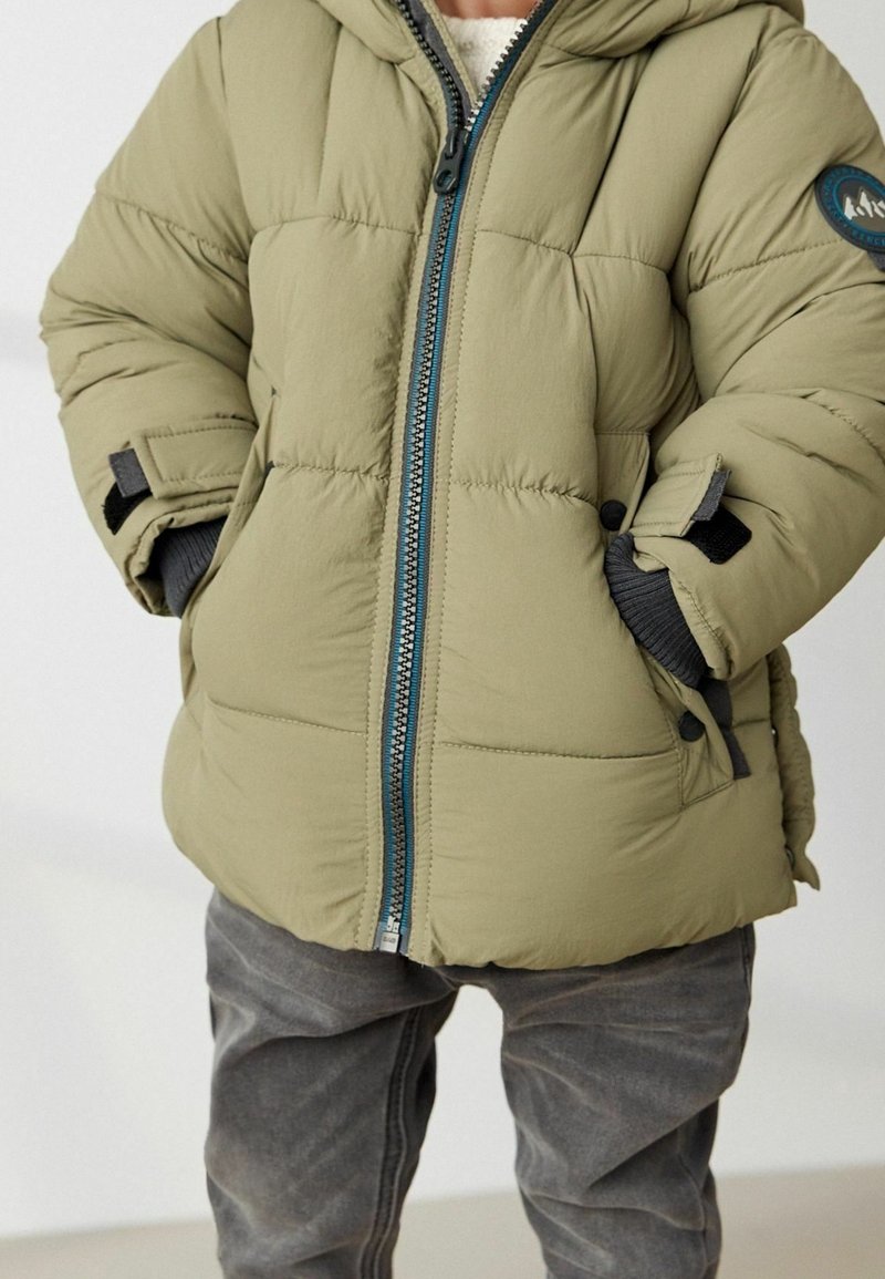 Chaqueta acolchada verde oliva con cierre de cremallera en la parte delantera, diseño acolchado, capucha y detalles en gris. Cuenta con un parche de logo redondo y puños ajustables.