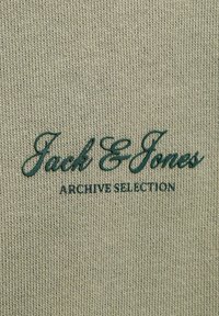 Texte vert « Jack & Jones Archive Selection » brodé sur un tissu tricoté beige.